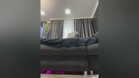 A video of a person lying on a gray sofa with a blanket, in a room with gray curtains and a window with greenery outside.