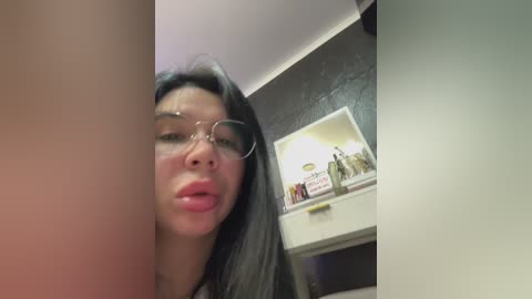 Video of an Asian woman with long black hair and glasses, making a kissy face, in a dimly lit room with a mirror and framed artwork on the wall.