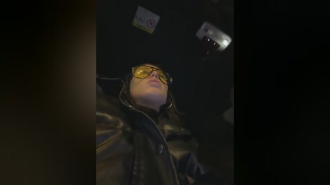 A video of a person in a dimly lit room, wearing a black leather jacket and yellow-tinted aviator glasses, looking upward with a neutral expression.