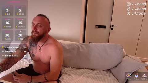 Video of a shirtless, muscular, bearded man with tattoos, sitting on a bed, holding a phone, smiling. Background shows a modern bedroom with light-colored walls and a closed wardrobe.