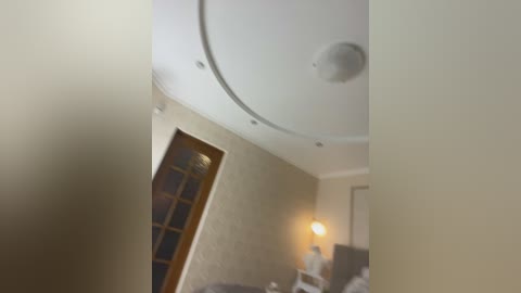 A blurry video of a bedroom ceiling with a circular light fixture and a wall-mounted lamp. The background includes a wooden door with glass panes and a bed with white sheets.