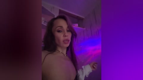 Video of a topless woman with medium skin tone, long dark hair, and a fit physique, standing in a dimly lit room with purple and blue lighting. She's wearing a white headband and necklace.