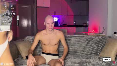 A photo of a shirtless, muscular, bald, white man in white underwear sitting on a gray, textured sofa in a dimly lit, modern kitchen.