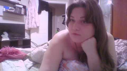 A candid video of a young, fair-skinned woman with long brown hair, lying on a bed with patterned sheets, in a messy bedroom.