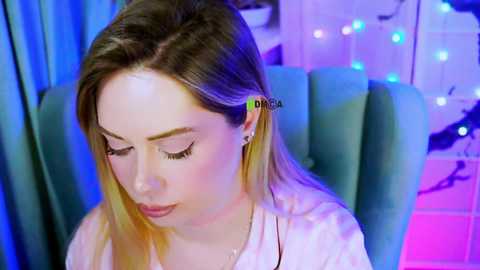 Video of a young woman with long blonde hair, wearing a white shirt, sitting in a teal chair. She has a headphone with a pink tip on her left ear. The background features a blue curtain and colorful string lights.