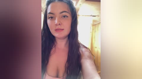 A video of a young woman with long, dark hair, fair skin, and a neutral expression, wearing a strapless top, taken indoors with soft lighting.