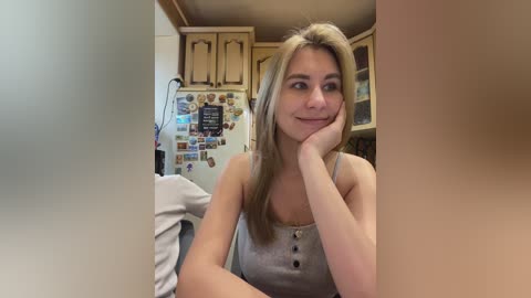 Video of a young, blonde Caucasian woman with fair skin, wearing a light gray tank top, sitting in a cozy, cluttered kitchen with wooden cabinets and a refrigerator adorned with magnets.