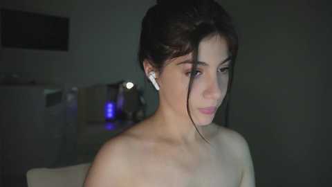 Video of a young woman with olive skin, dark hair tied back, wearing white earbuds, topless, in a dimly lit room with blurred background.