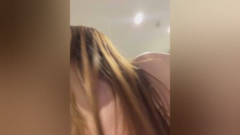 A video captures a close-up of a woman's long, straight brown hair partially obscuring her face, with her head tilted downwards. The background is blurry, featuring a beige wall and a ceiling light.