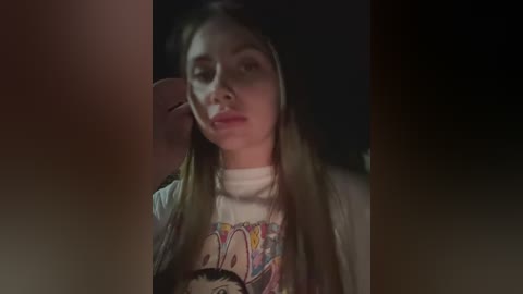 Video of a young woman with long blonde hair, wearing a cartoon-print t-shirt, sitting in a dimly lit room.