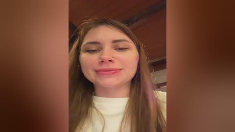 Video of a young Caucasian woman with straight, light brown hair, fair skin, and closed eyes. She wears a white top. Background shows wooden ceiling and blurry walls.