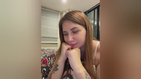 Video of a fair-skinned woman with long, straight brown hair, wearing a floral dress, resting her chin on her hands, in a modern bedroom with white walls and a floral-patterned bedspread.