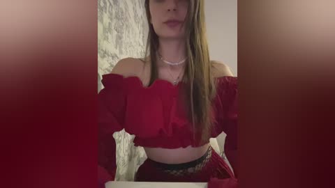 Video of a young woman with long brown hair, wearing a red off-the-shoulder crop top and a patterned skirt, standing in front of a textured wall, partially obscured by soft lighting.