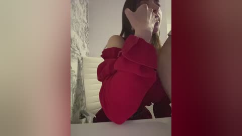 Video of a woman with long, dark hair and fair skin, wearing a red off-shoulder dress, sitting on a white leather chair in a modern bathroom with a tiled wall.