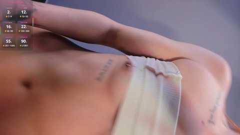 Video of a fair-skinned woman with a toned, flat stomach, wearing white panties, with a tattoo of a quote on her left ribcage. Background shows a blurred, out-of-focus room with a time display.
