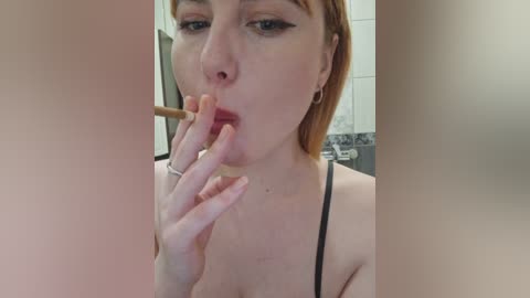Video of a fair-skinned woman with short red hair, wearing a black strapless top, sucking on a wooden stick. Background shows a tiled bathroom with a shower curtain.