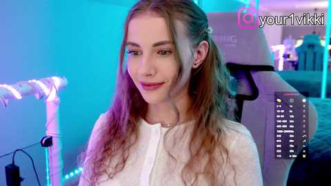 Video of a smiling, light-skinned woman with long, wavy brown hair, wearing a white top, sitting in a gaming chair in a brightly lit, modern room with neon lights.