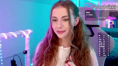 Video of a young, fair-skinned woman with long, wavy brown hair, wearing a white top, smiling softly. Background features a gaming setup with neon lights, and a chat window displaying her username \"@yourvikki.\