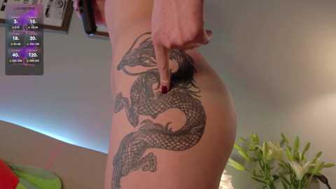 Video of a woman with fair skin, showing off a detailed black dragon tattoo on her left hip. She is standing in a modern, minimalist bedroom with a green plant and a bed visible in the background.
