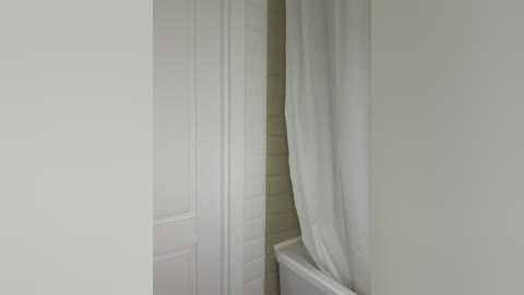 A video of a minimalist bathroom corner with white walls, a white door slightly ajar, and a white curtain partially drawn, revealing a white bathtub. The scene is simple and clean, emphasizing a serene, uncluttered ambiance.