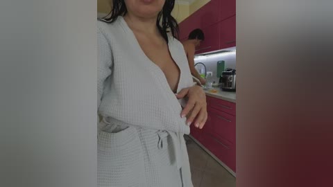 Video of a woman in a bathrobe, standing in a modern kitchen with red cabinets and stainless steel appliances. Her hand is on her breast, partially revealing her nipple.