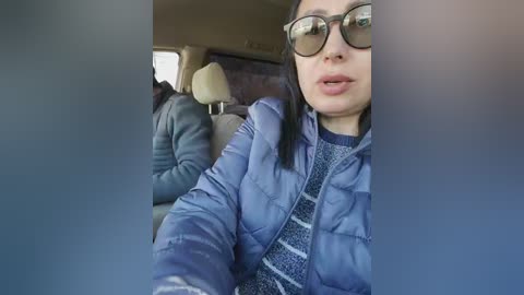 Video of an Asian woman with glasses, wearing a blue puffy jacket and a striped sweater, seated in a car with beige seats.