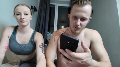 Video of a fit, shirtless man with short brown hair and a tattoo on his shoulder, holding a black smartphone. Next to him, a woman with fair skin, brown hair, and a grey sports bra, holding a pink bandage. Both indoors, near a door.