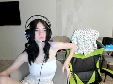 A video of a young woman with long black hair, wearing a white tank top and headphones, sitting in a modern living room with a flat-screen TV, a green chair, and a white-patterned couch.