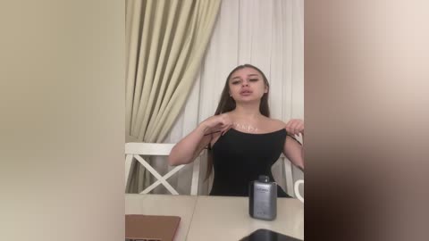 Video of a young woman with long brown hair, light skin, and a slender physique, wearing a black tank top, adjusting it, seated at a table with a bottle of lotion. Background features beige curtains and a white chair.