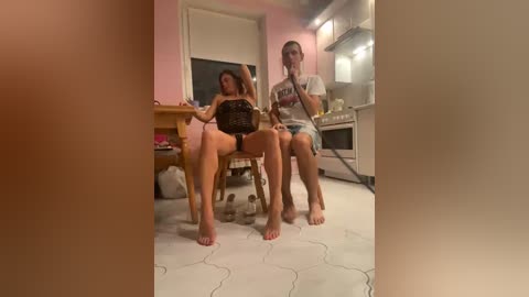 Video of a woman with brown hair, wearing a black top, sitting barefoot on a chair in a small kitchen with a beige floor, another woman in a white shirt and denim shorts standing behind her.