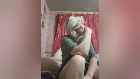 Video of a middle-aged, short-haired woman in a green T-shirt, sitting on a bed with red curtains and floral wallpaper in the background. She rests her chin on her hand, appearing thoughtful.