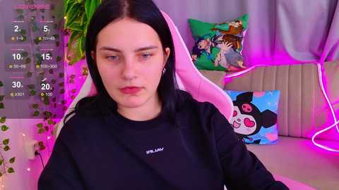 Video of a young woman with long black hair, wearing a black sweatshirt, seated in a pink gaming chair in a room with vibrant neon lights, colorful pillows, and green plants.