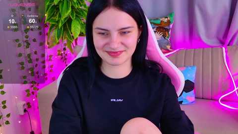 Video of a young woman with long black hair and fair skin, wearing a black shirt, sitting in a gaming chair with pink accents, surrounded by vibrant, neon-lit decor and green plants.