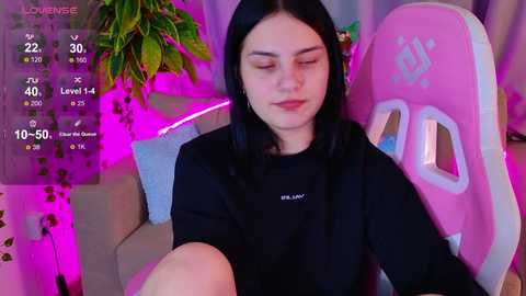 Video of a young woman with long black hair, wearing a black shirt, sitting on a pink gaming chair with green accents. Background includes purple lighting, potted plants, and a digital overlay with game statistics.