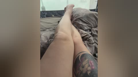 Video of a person lying on a bed, showcasing their tattooed thigh and bare legs, with a messy brown bedspread in the background.