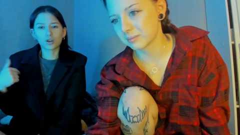 Video of two women in a dimly lit room. One wears a black coat and talks animatedly, while the other, with tattoos, dons a red plaid shirt, looking contemplative.