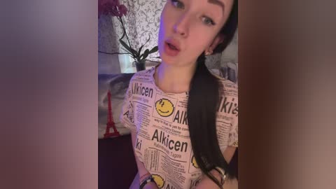 Video of a young woman with light skin, long dark hair, and wearing a pink \"Alki\" t-shirt. She's in a dimly lit room with a red Eiffel Tower figurine and a potted plant.