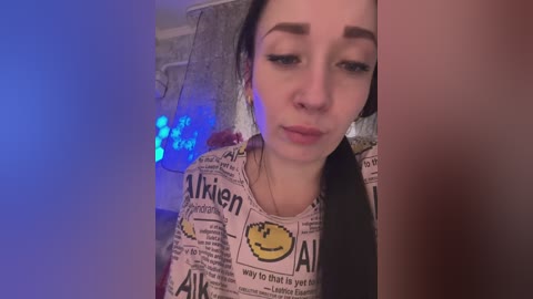 A video of a young woman with long black hair, fair skin, and a small nose, wearing a Alien-themed T-shirt. Background features a blurred, dimly lit room with blue lights.