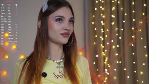 Video of a young woman with long brown hair, light skin, and blue eyes, wearing a yellow shirt, silver hoop earrings, and a cross necklace. Background features bokeh lights and a calendar.