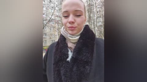 A video of a light-skinned woman with a bald head, wearing a black fur scarf, beige scarf, and dark coat, standing in a winter city with bare trees and beige apartment buildings.