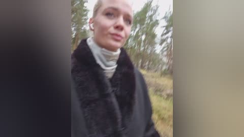 A video of a fair-skinned, bald woman with light makeup, wearing a black fur coat over a white turtleneck, standing outdoors in a blurred forest background.