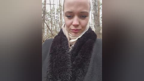 Video of a bald woman with fair skin, wearing a dark fur scarf and light scarf, against a backdrop of bare trees in a forest.