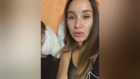 Video of a young woman with long, light brown hair and fair skin, wearing a black sleeveless top, lying on a white bed with a pillow. Background shows a tan wall.