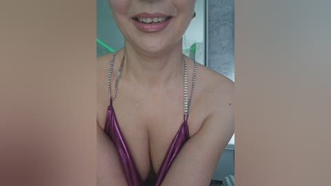 Video of a smiling woman with light skin, wearing a shiny, metallic pink halter top that accentuates her cleavage, with a blurred background featuring a green plant and a mirror.