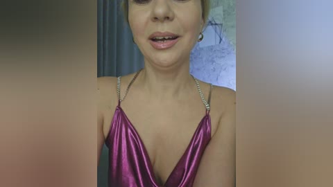 Video of a light-skinned woman with short blonde hair, wearing a shiny pink halter dress with thin straps, smiling with visible teeth, indoors with a blurred, textured wall in the background.