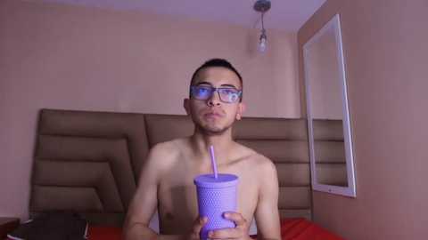 Video of a shirtless young Asian man with glasses and a mustache, holding a purple straw cup, standing in a dimly lit room with beige walls and a brown padded headboard.