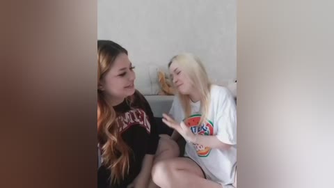 A video of two young women, one with long, wavy brown hair in a black shirt, and the other with straight blonde hair in a white shirt, sitting closely on a beige sofa, smiling and interacting warmly.