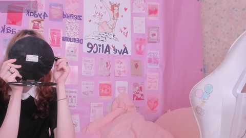 Video of a young woman with long, wavy red hair wearing a black mask, standing in a pink room adorned with posters and a white dollhouse.