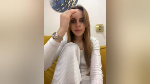 Video of a young Caucasian woman with long, straight brown hair, wearing white ribbed leggings and a white top, sitting on a mustard-yellow couch, touching her forehead, against a beige wall with a gold light switch and a blue and gold abstract painting.