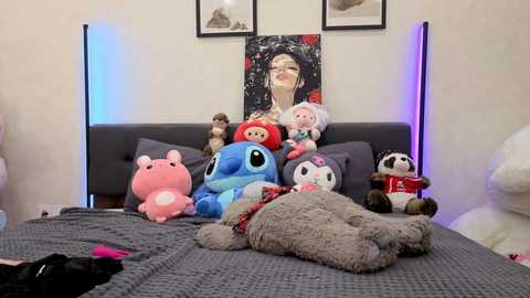 Video of a cozy bedroom with a plush gray bedspread, featuring a stuffed Snoopy, a red-headed Snoopy, a blue Stitch, and a white panda, surrounded by framed artwork and soft lighting.
